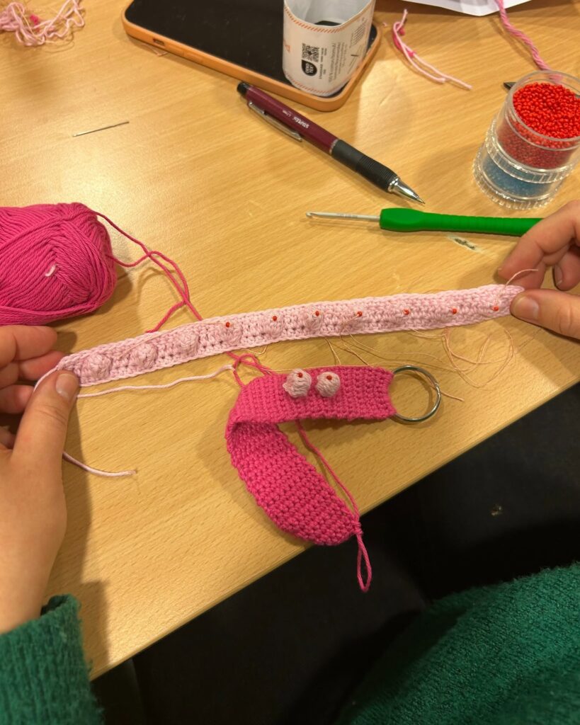 Ljusrosa virkat band med röda pärlor hålls upp; ett färdigt rosa band med nyckelring ligger bredvid