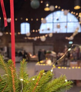 Grangrenar med ljusslingor i förgrunden och en julmarknad i en upplyst lokal i bakgrunden