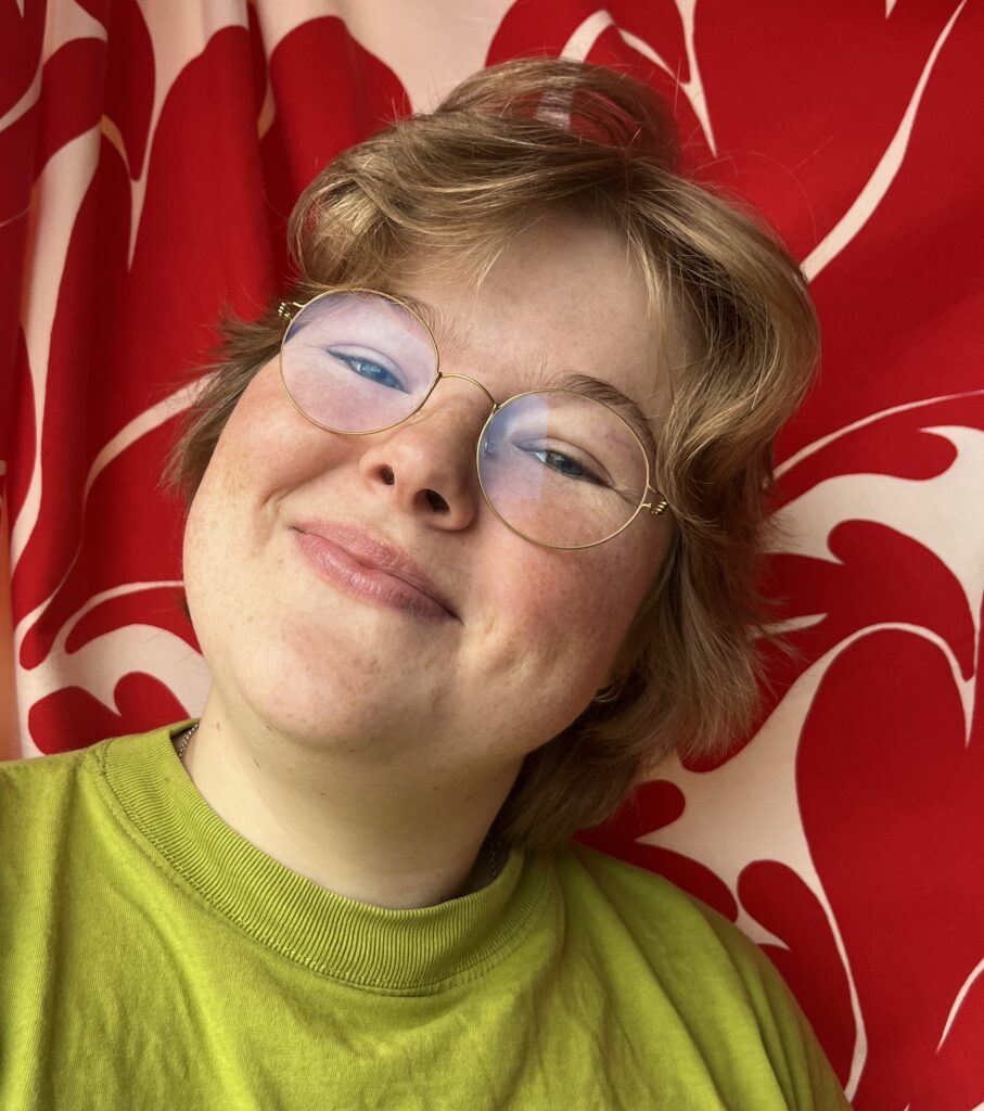 Ebba Hernström med glasögon och grönt plagg framför ett röd- och vitmönstrat tyg