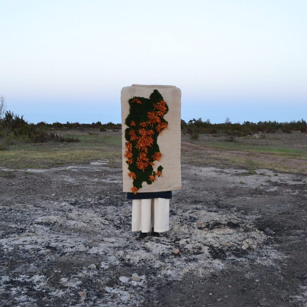 Person i vitt står i ett kargt landskap bakom en handgjord textil med grönt och orange blomsterbroderi