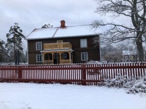 Ett mörkbrunt trähus med snötäckt tak och veranda i Stensjö by, omgivet av snöiga träd