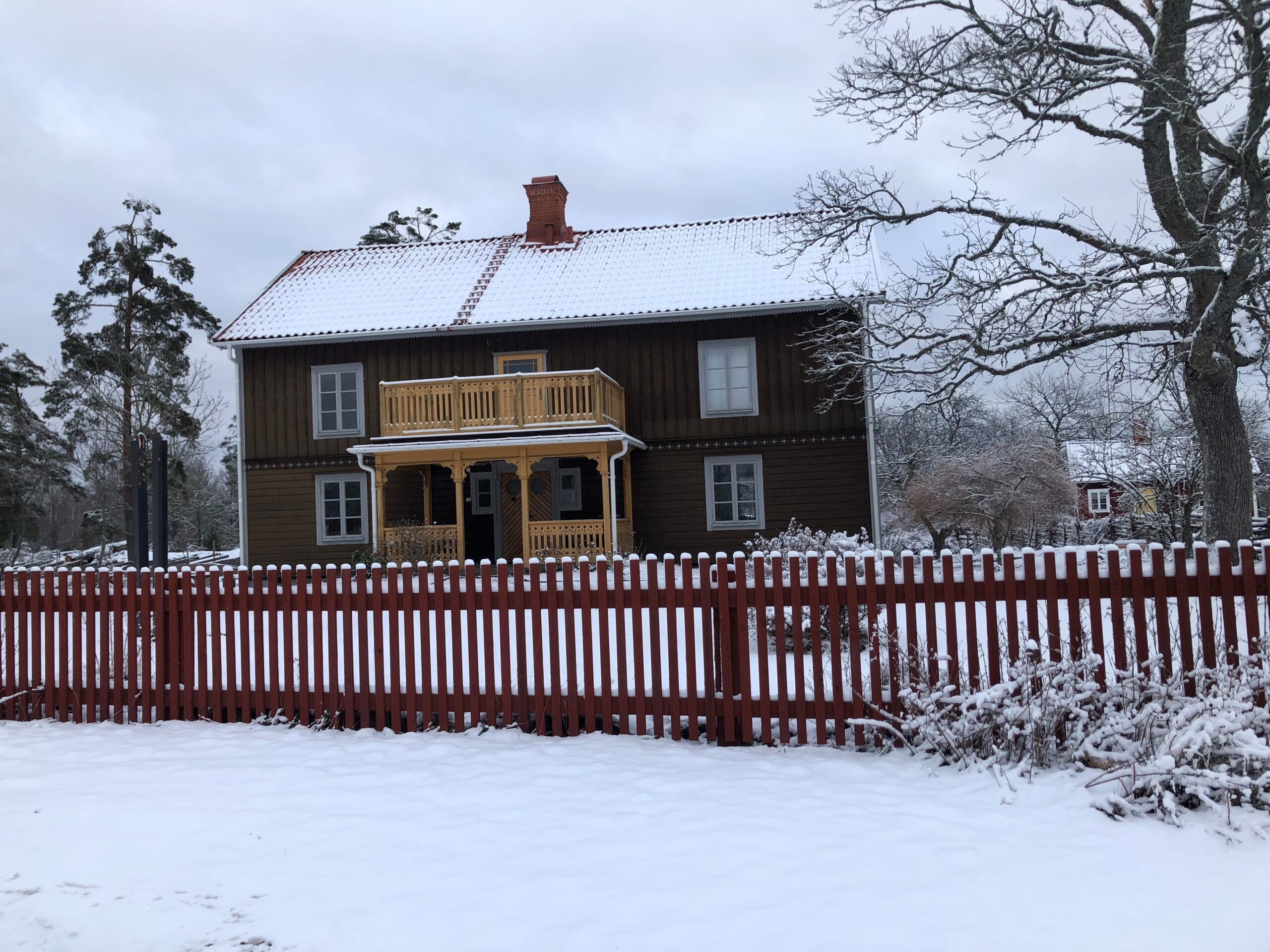 Ett mörkbrunt trähus med snötäckt tak och veranda i Stensjö by, omgivet av snöiga träd