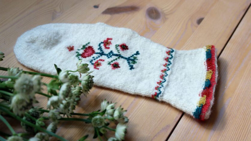 Vitt handbroderat ylleföremål med färgglada blommor och bårder, på ett träbord bredvid blommor