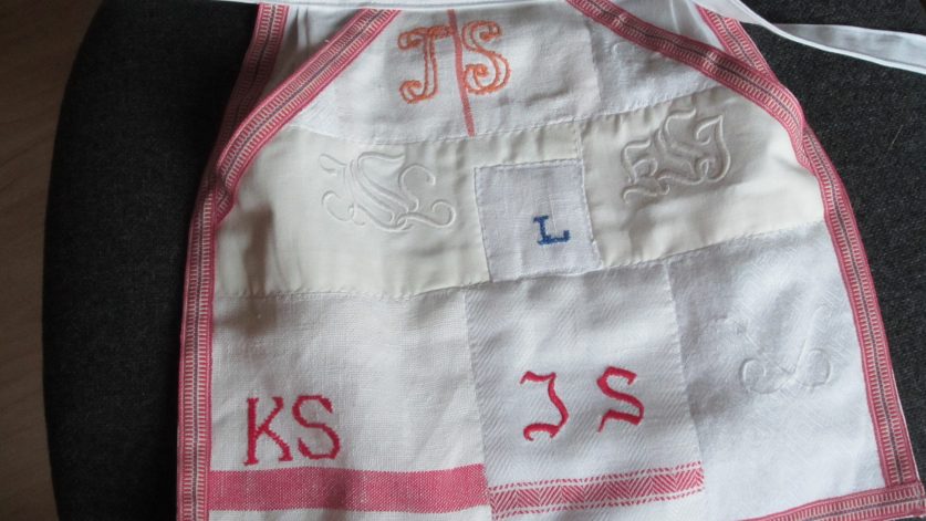 Litet förkläde i rött och vitt med olika initialer: JS, KS, L.