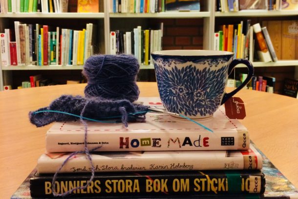 Mörklila garnnystan och blå, blomstermönstrad tekopp står på en hög med böcker, staplade på bord framför en bokhylla.