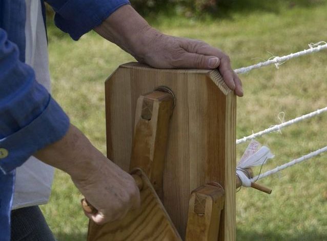 Repslagningsmaskin tillverkad i träd, med vev och tre hållare att fästa material i. En person står utomhus och tvinnar rep av trasor.
