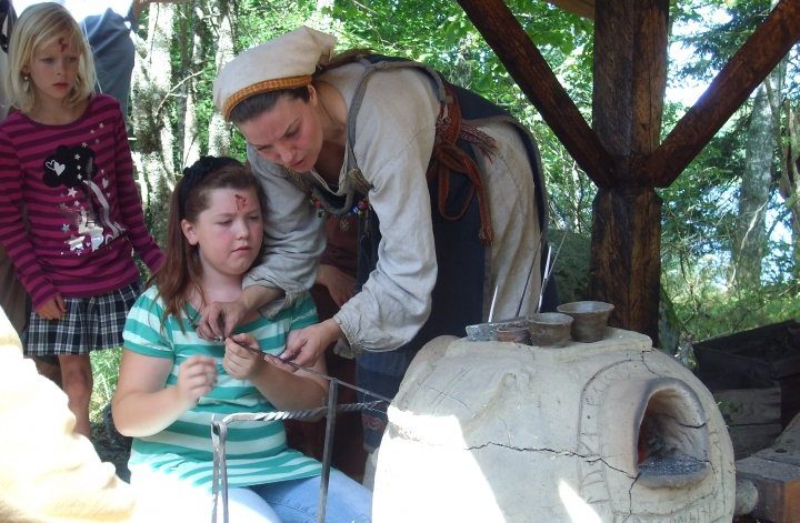 EN kvinna klädd i vikingatida kläder visar ett barn hur man gör pärlor